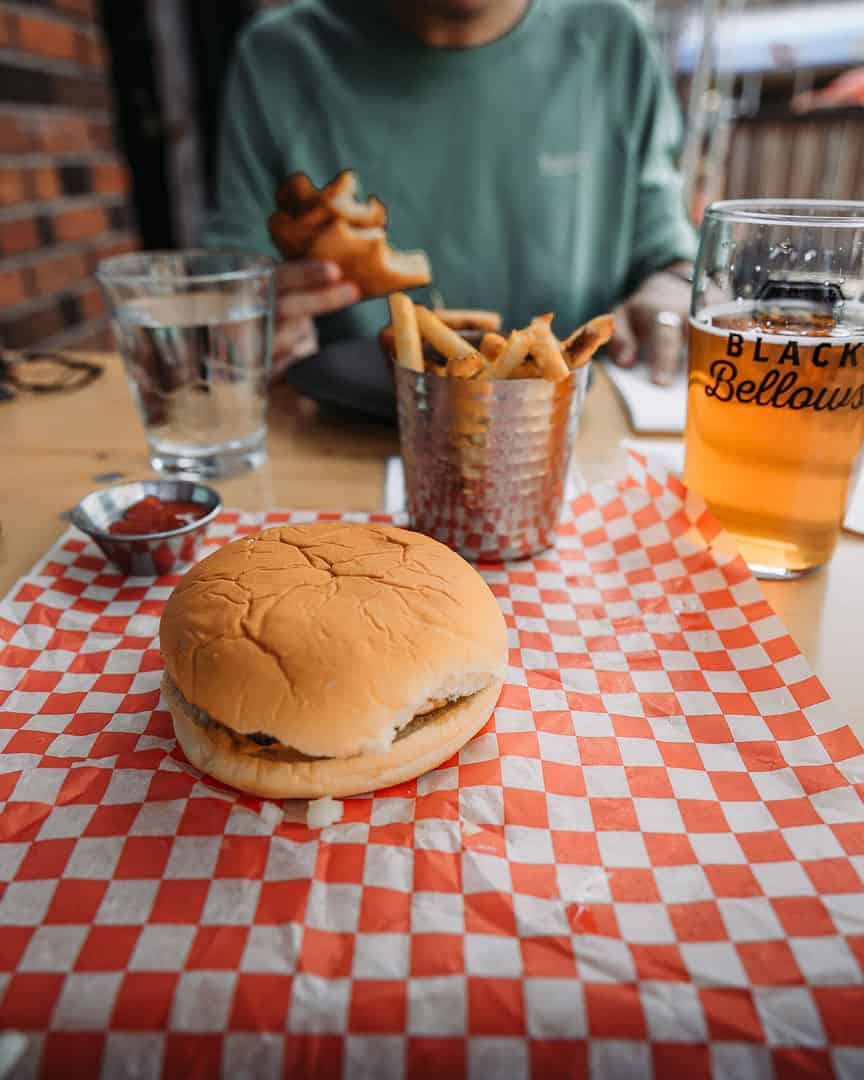 the photo is from the view of someone sitting at a table at a restaurant. in front of them is a burger, fries and beer on the table. the burger is on a red and white checkered sheet. the person across from them is wearing a light green shirt. you do not see their head. the foreground is the table and burger. the background is the person