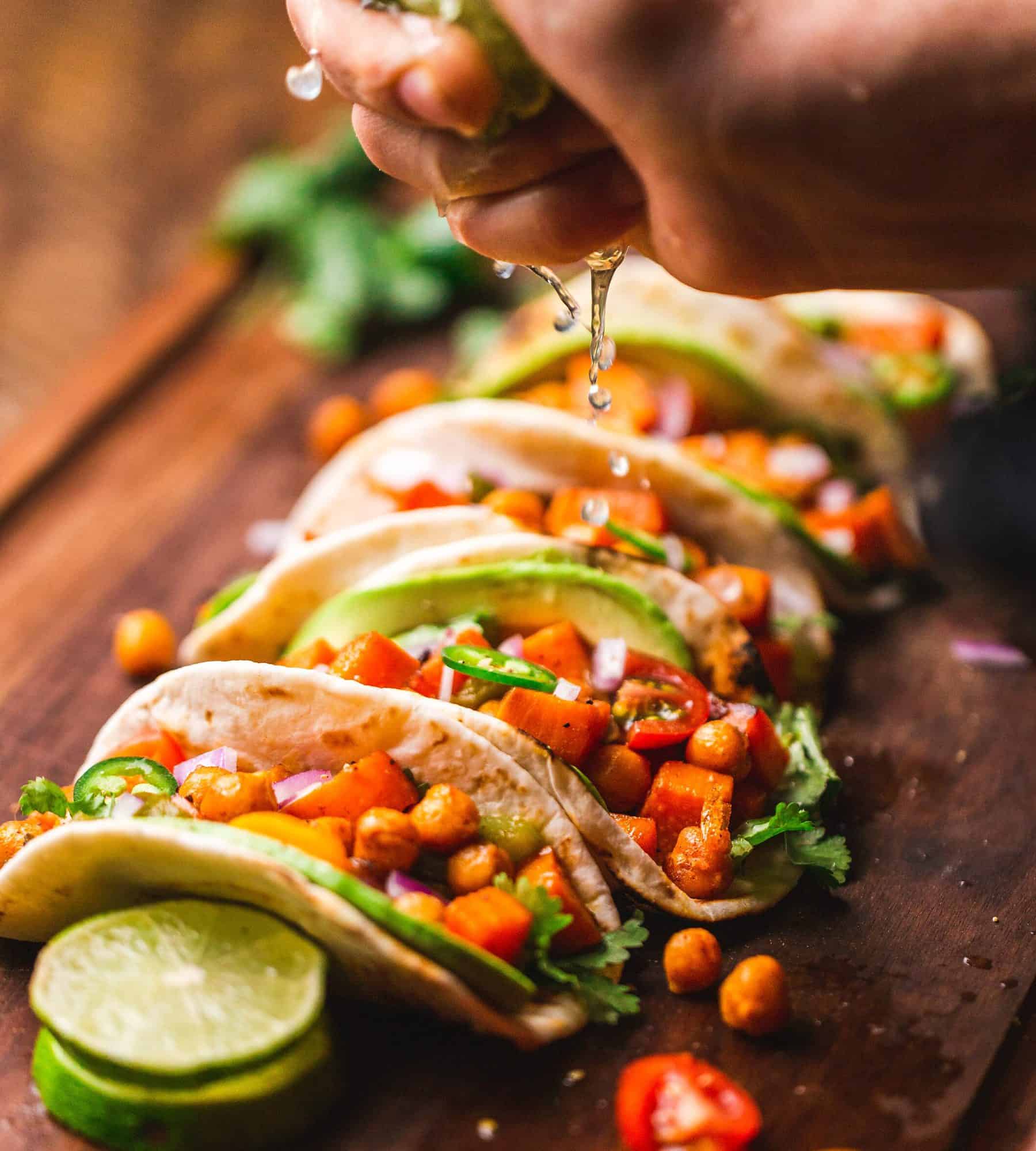 photo shows a hand squeezing a lime over multiple tacos filled with veggies. the background is a blurred wooden table. the foreground is the hand and tacos