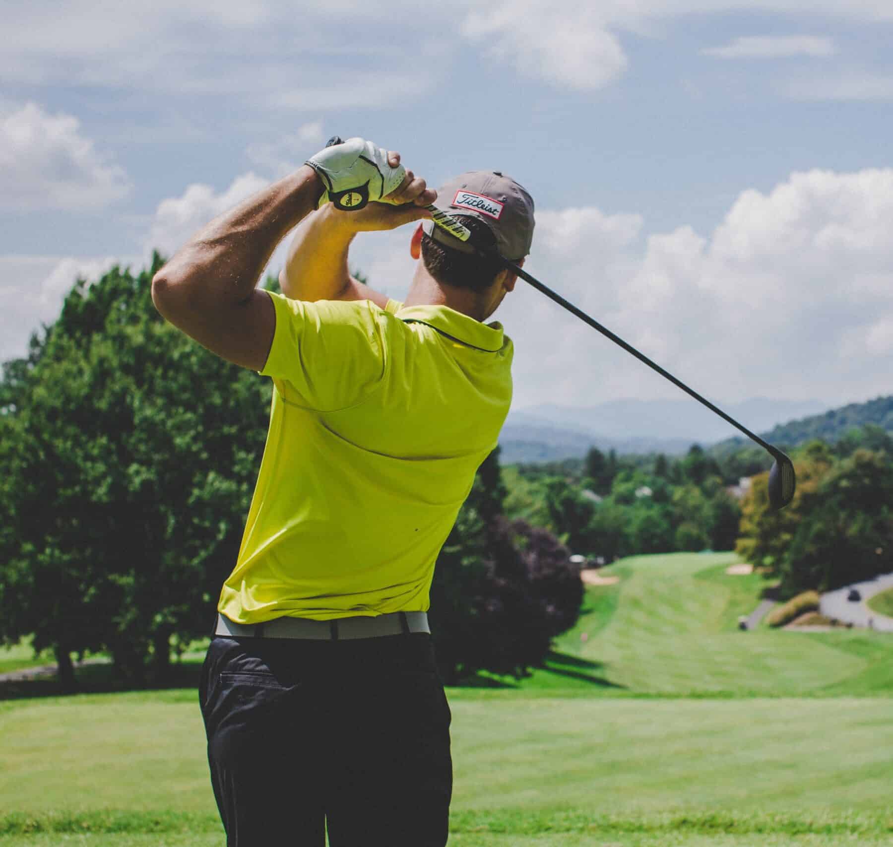 a man is playing golf in a bright green t-shirt. he is in the swinging position. it is a medium shot with his body cut off just above his knees. the foreground is the man. the background is a beautiful golf course on a part sunny day with white clouds