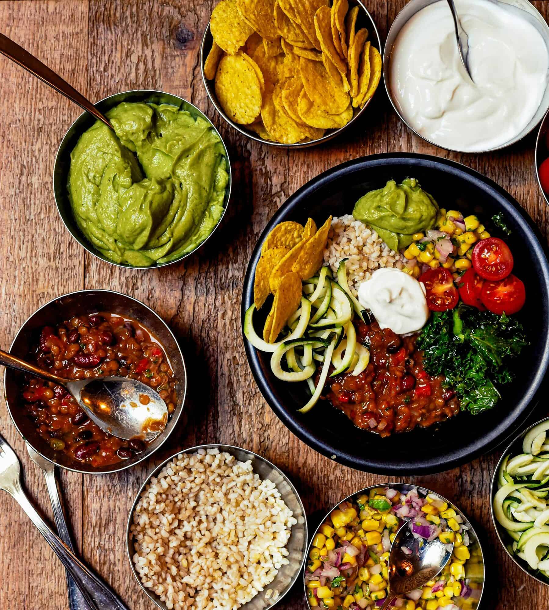 one large black bowl is in the middle of 9 smaller bowls surrounding it. each small bowl has different Mexican ingredients such as chips, guac, tomatoes, salsa, greens, sour cream and peppers. the background is a brown table. the foreground is the bowls of food. the angle of the shot is looking down from an aerial view.