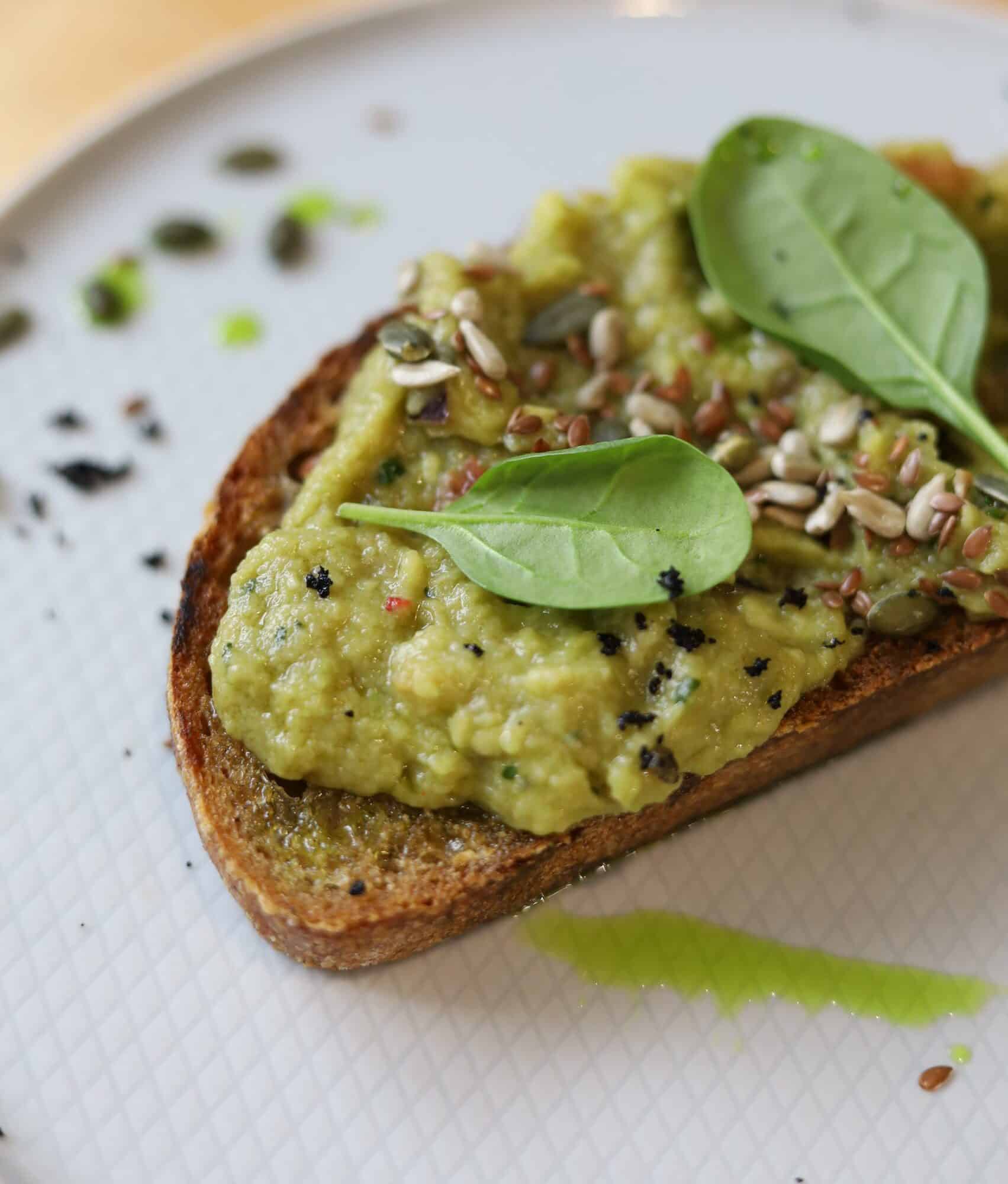 avocado toast made with avocado, sunflower seeds and spinach is sitting on a white plate. there is one slice of the toast. the background is the white plate and a hint of a light brown table. the foreground is the avocado toast