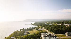 the photo is an aerial view of south georgian bay. the rolling hills are in the background and you can see the shoreline of the bay. the grass is green and trees are bloomed. there are some buildings in the shot but it is mostly greenery and the shoreline