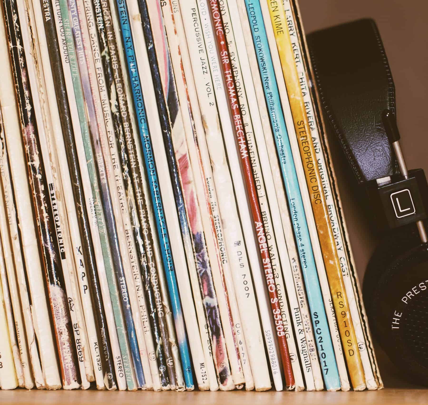the image shows a stack of vintage vinyl records sitting upright on a brown shelf. there is a pair of over ear black headphones on the right of them.