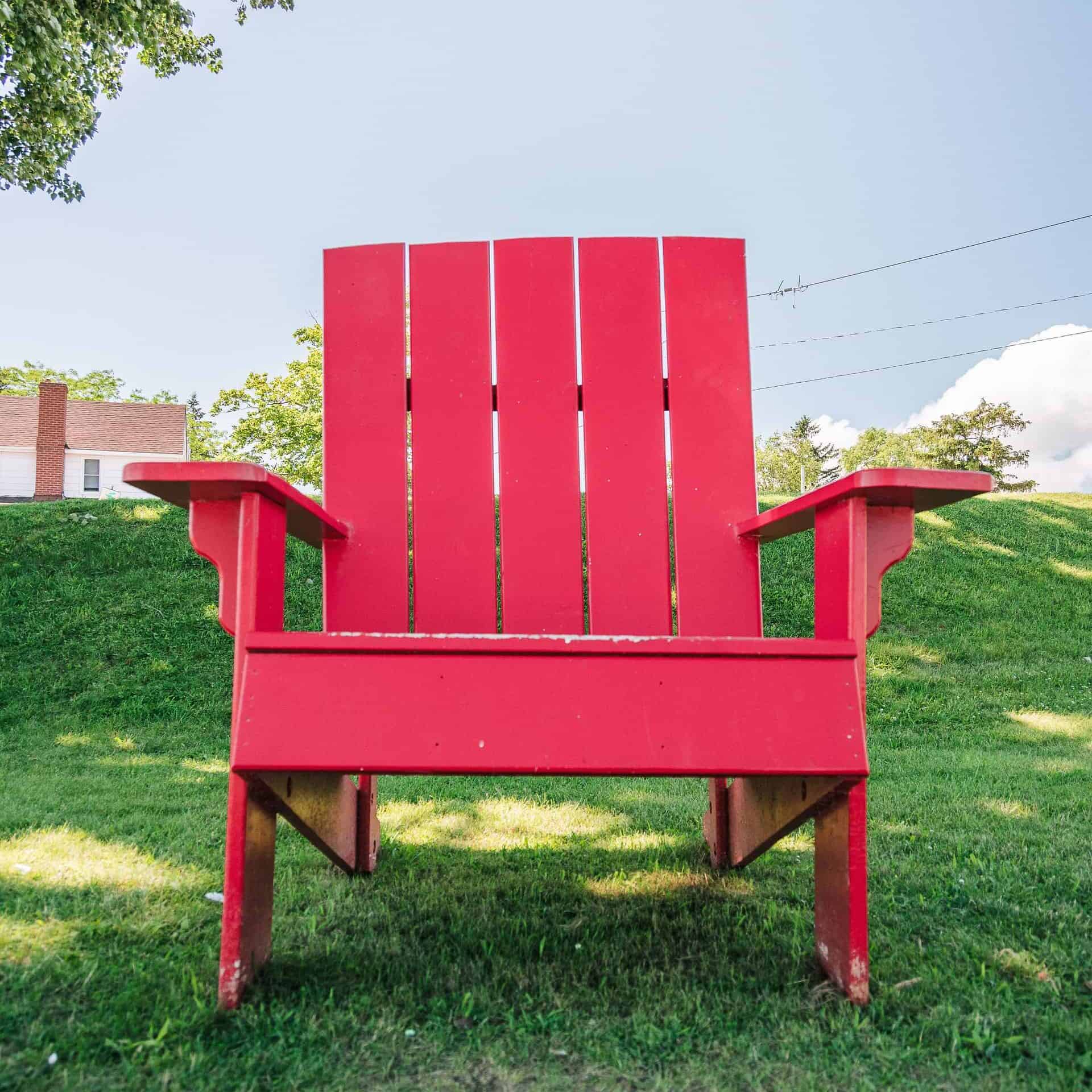 a big red chair is sitting on the green grass on a beautiful sunny day. the chair is the style of a muskoka chair.