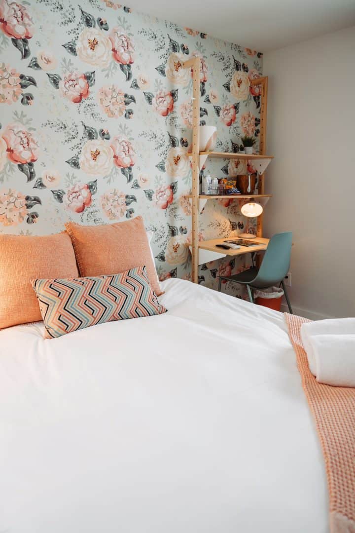 this room has a white bed in it with light peachy pillows. the wall paper is light blue with pastel flowers on it. i nthe corner there is a small light desk to do some work at
