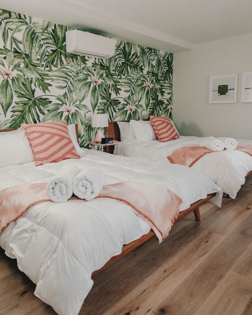 2 beds in a retro 70s inspired room. there is white and light pink bedding and palm tree wall paper on the walls